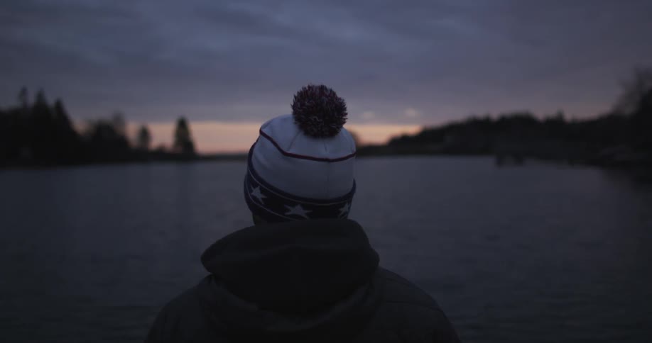 A man in a winter knit cap stands looking across a lake at dusk