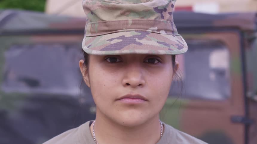 A woman standing and staring with a military uniform with a vehicle in the background
