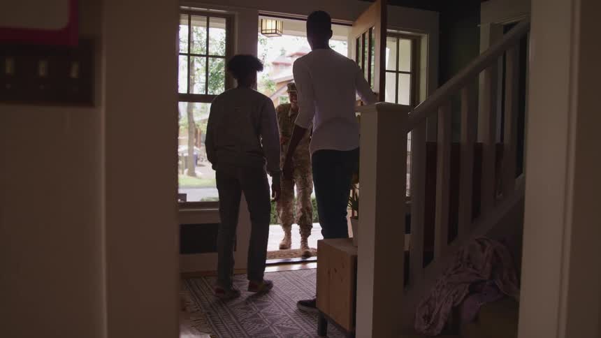 A man opening the front door of a house and hugging a person with a military uniform