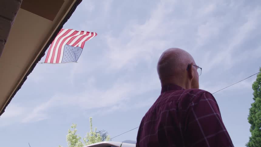 Elderly man standing outside his house under an american flag waving in the wind