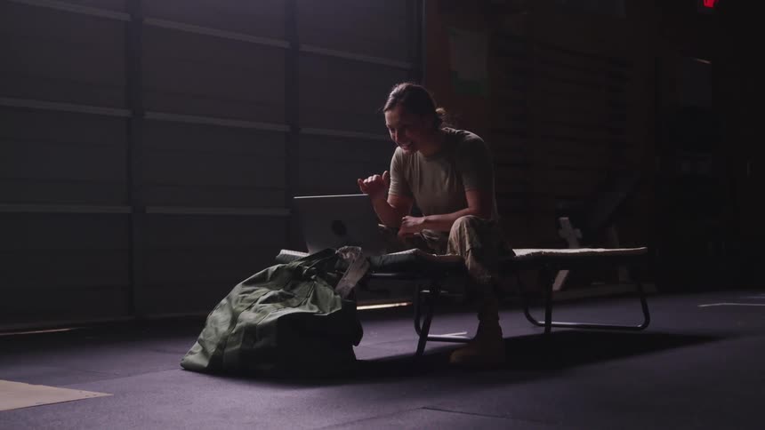 Young female army soldier sitting on a bed italking on a video call on a laptop