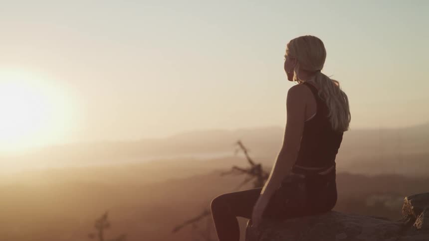 A teenage girl sits on a rock on a cliff and looks out at the landscape
