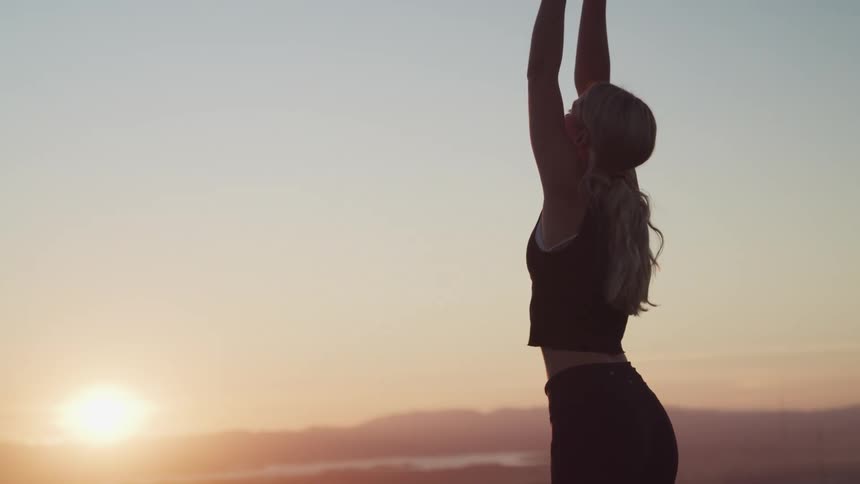 The silhouette of a young woman can be seen as she stretches her arms upward while exercising outdoors