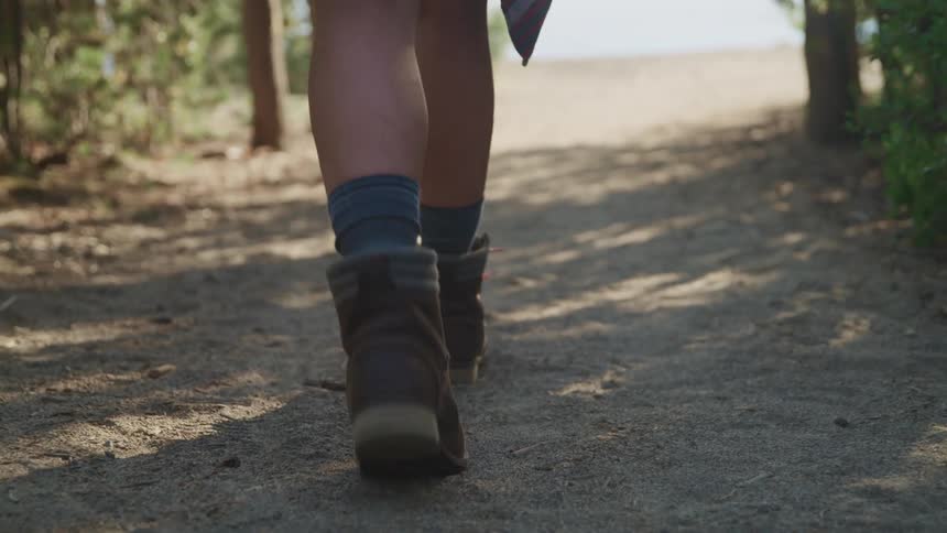 Woman wearing boots walks on a dirt trail through the forest at evening