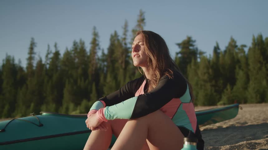 A woman sitting on the ground with a kayak trees and the sky in the background