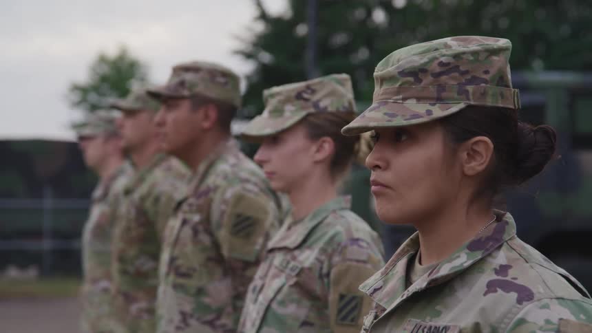 Uniformed soldiers stand side by side in a line near armored vehicles