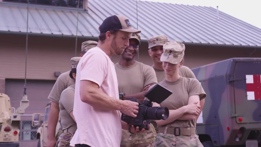 Soldiers in a military base looking at a camera with vehicles buildings sky and clouds in the background
