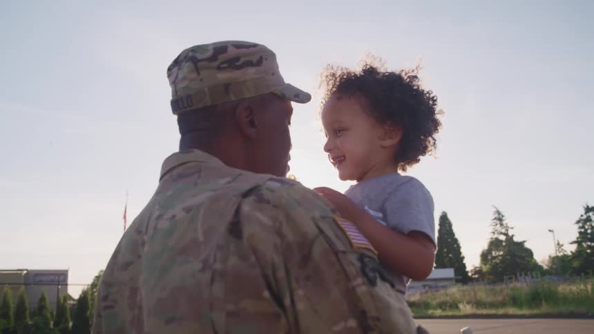 A uniformed soldier holds his son outdoors and gives him a kiss