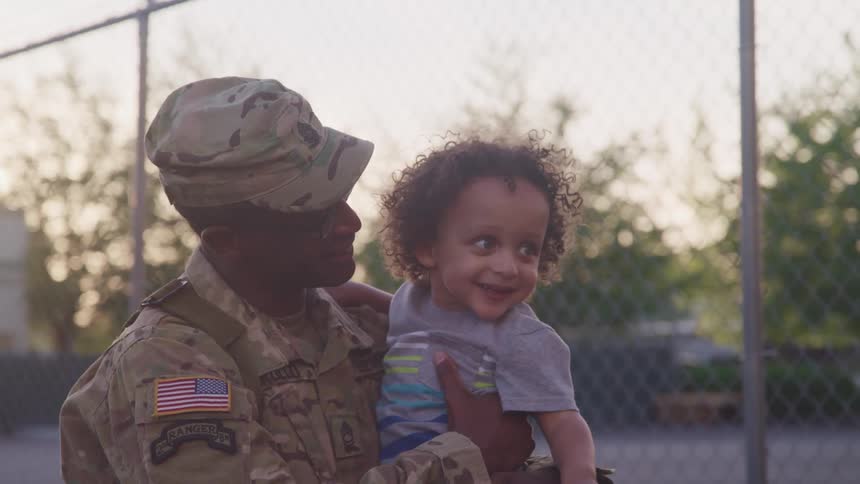 Father wearing fatigue holds child while sitting near chain link fence