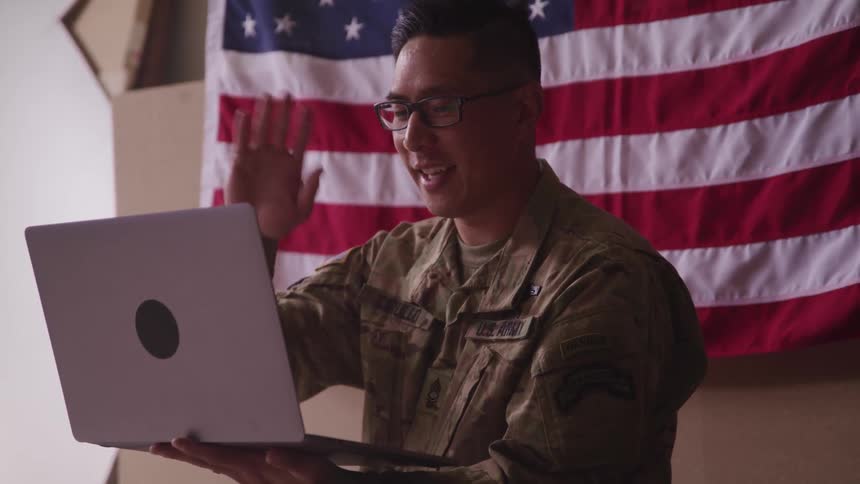 A us army soldier sitting in front of an american flag waves at a laptop camera he holds in his hand and smiles