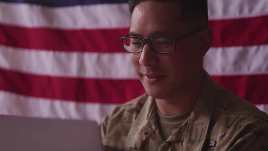 A young soldier sitting in front of an american flag waves at a laptop during a video call