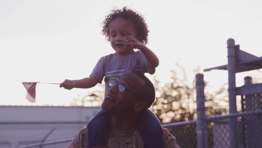 A soldier holds his young son on his shoulders as the toddler waves a small american flag
