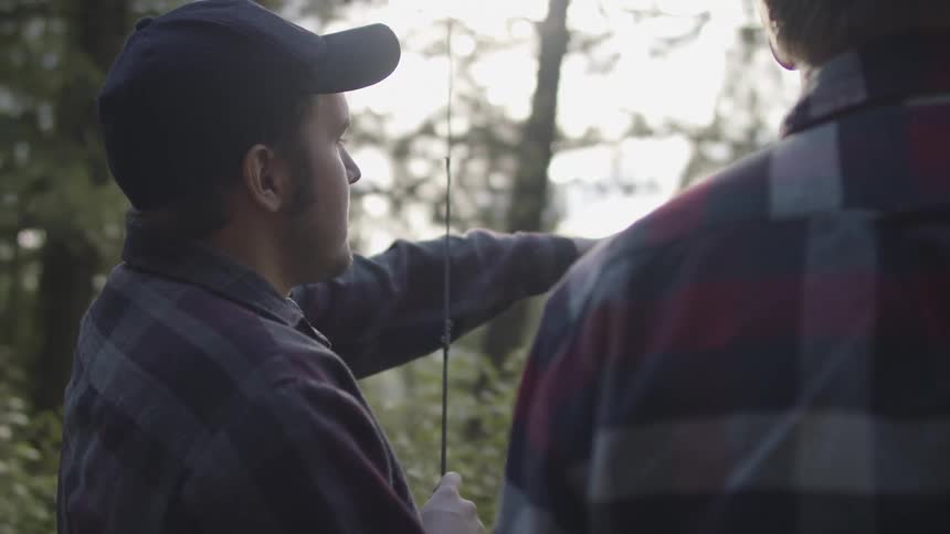 A man with a hat and a fishing rod talking to another man with trees the sky and clouds in the background