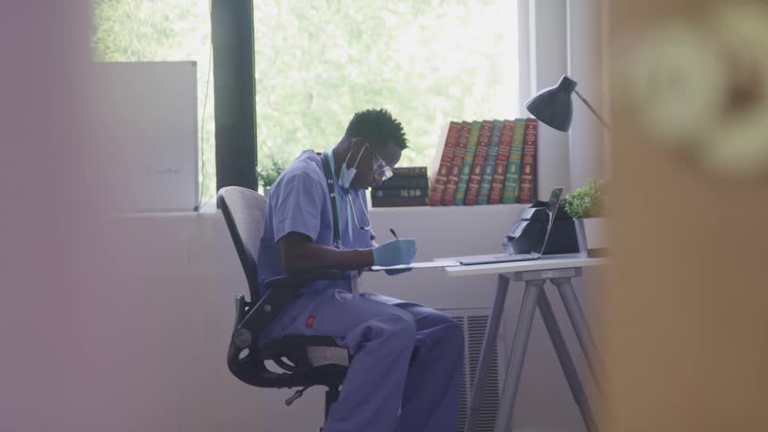 A male medical professional in scrubs sits at a desk in his office