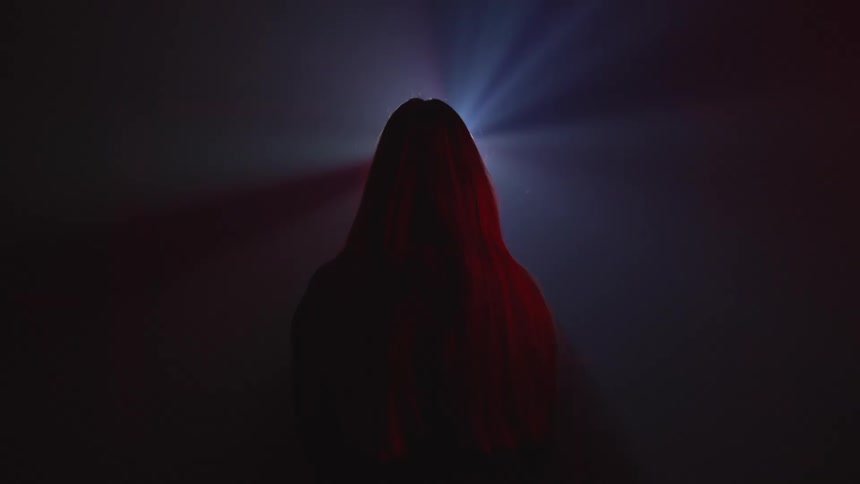 Long haired woman standing in front of the colorful moving lights of a spotlight over the smoke inside a dimly lit room