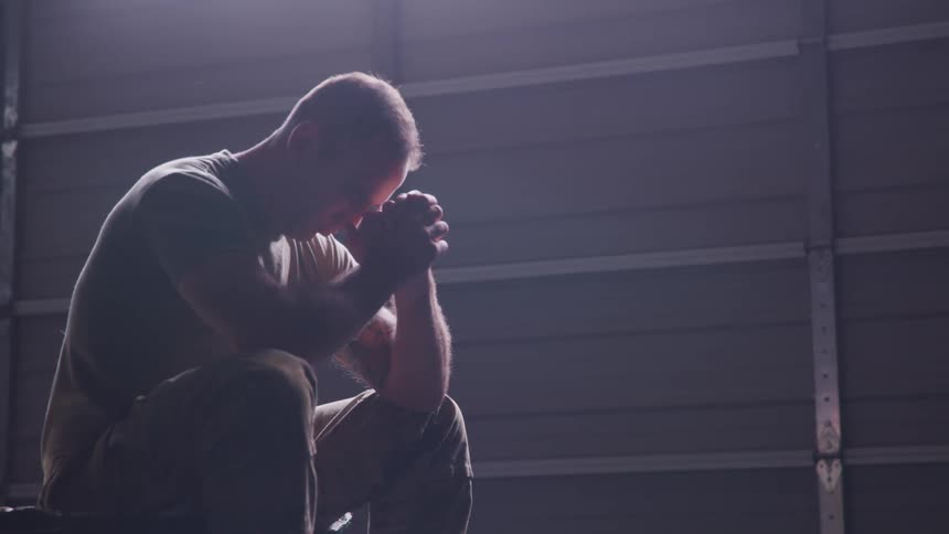 A depressed soldier sits contemplatively with his eyes closed before resting his head on his clenched hands