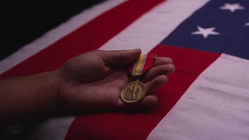 A person caresses a gold medal while standing with their arm resting on a casket draped with an american flag