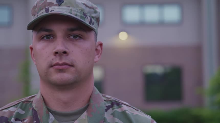 A soldier in a camouflage uniform stands in front of a brick building