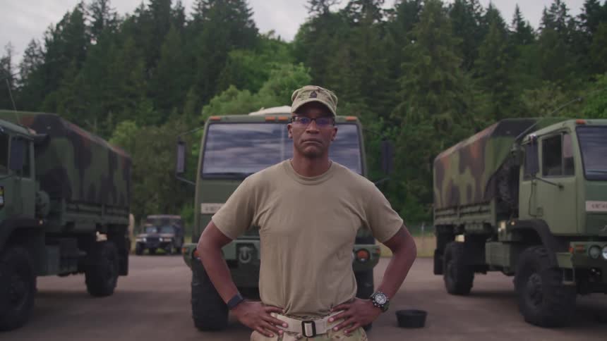 A soldier stands with his hands on his hips in front of camouflaged trucks