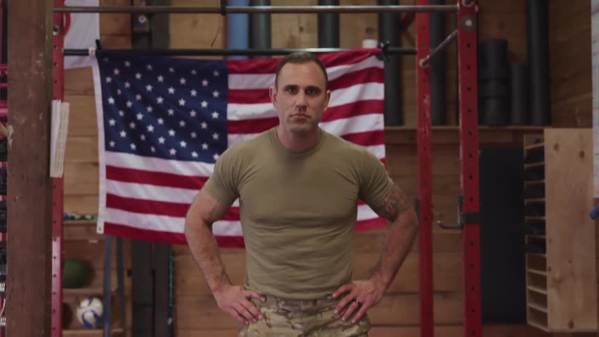Strong muscular american soldier standing in front of an american flag inside a room looks up at the camera
