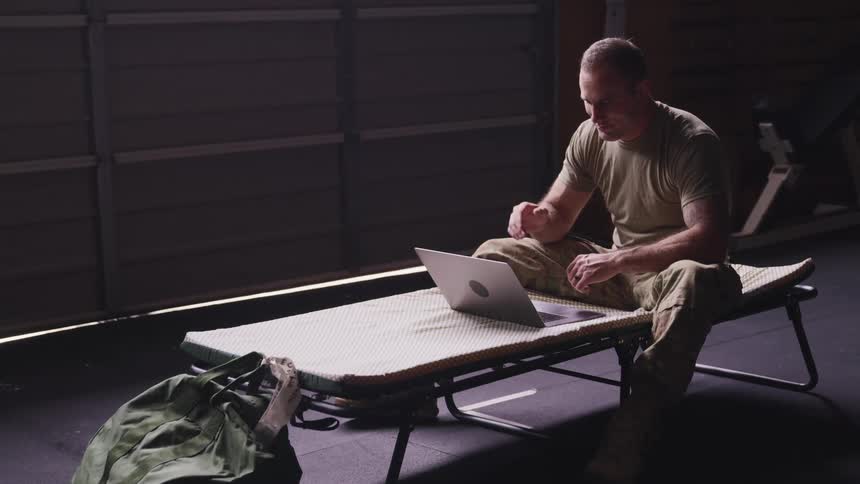 Soldier sittin in a bed on a dimly lit room waves goodbye on a video call through a laptop