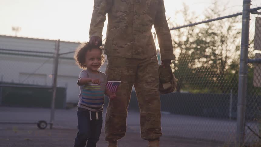 A toddler boy waves an american flag while walking with an adult wearing a military uniform