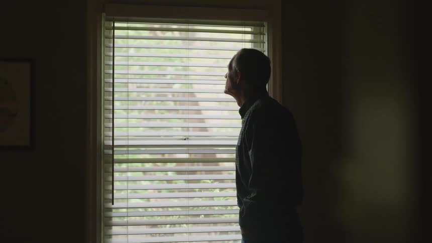 A man standing in a room next to a window stares outside