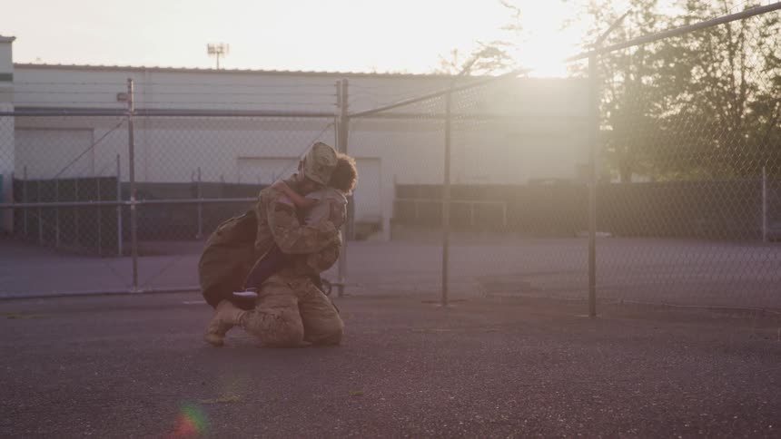 African american soldier kneeling on the ground to hug his son on a yard at sunset