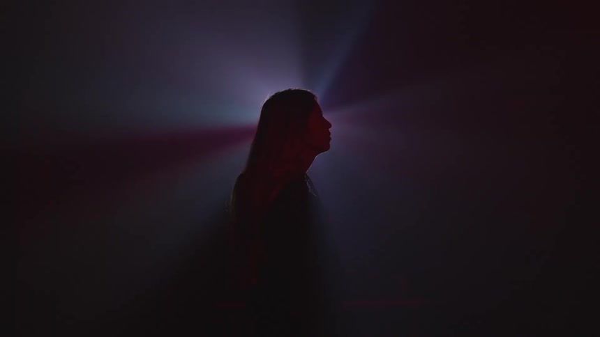Silhouette of a long haired woman stainding in front of the colorful lights of a spotlight in a smoke filled dark room