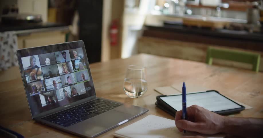 A man sitting at a table holding a pen and video chatting on a laptop