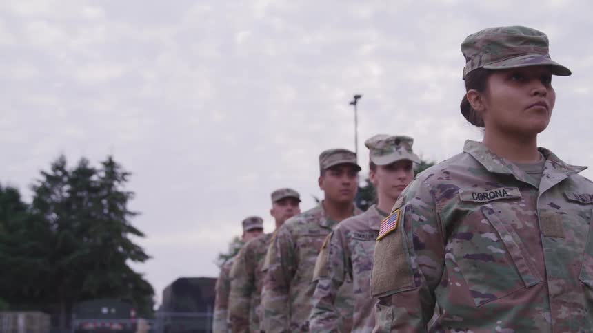 Soldiers in military uniforms lining up and turning with vehicles trees the sky and clouds in the background