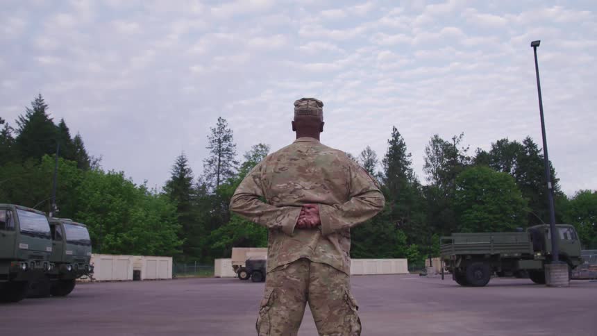 A soldier stands in front of army trucks with his hands clasped behind his back