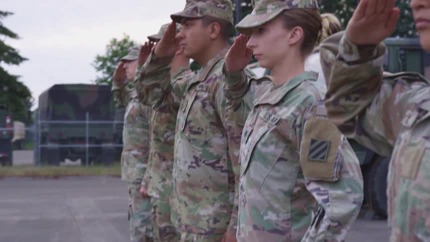 A group of soldiers salute while standing shoulder to shoulder in a military parking lot