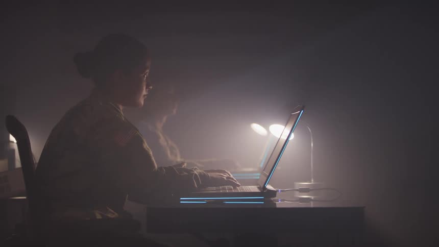Two young woman in military uniforms sitting at desks and typing on laptops while also looking up at something