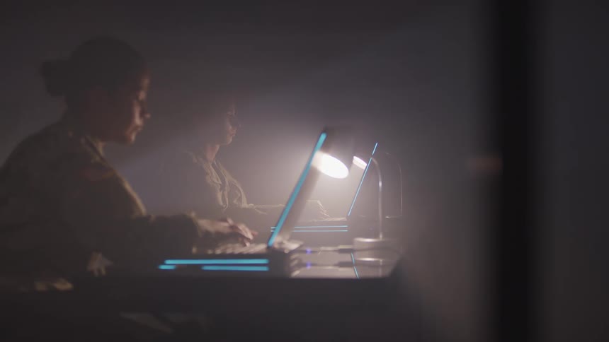 Two women with military uniforms sitting at desks typing on laptops