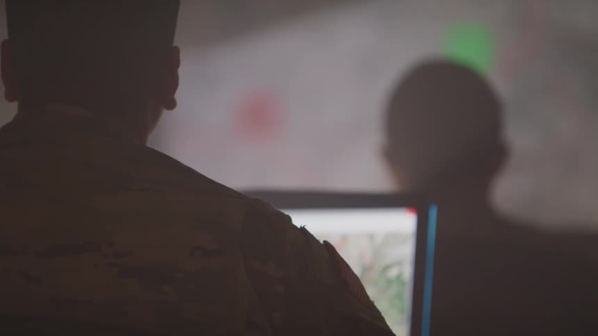 Two soldiers sitting and watching a presentation on a projector screen with laptops