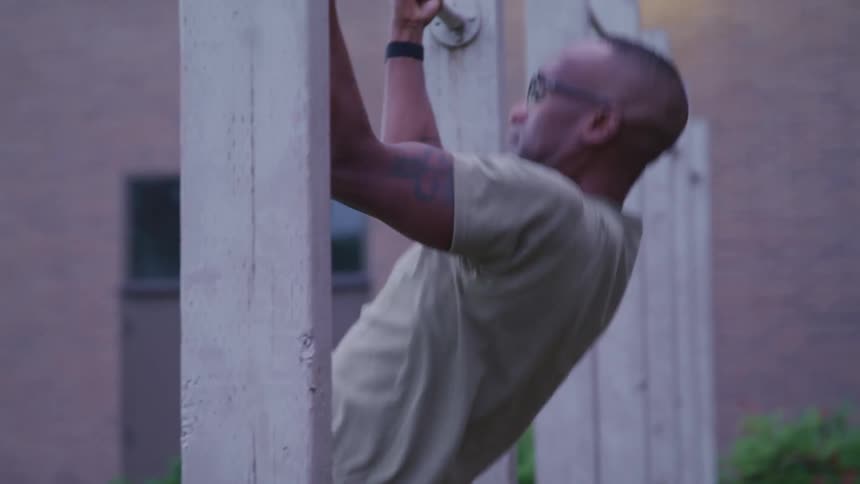 A man with a military uniform doing pull ups with a building and plants in the background