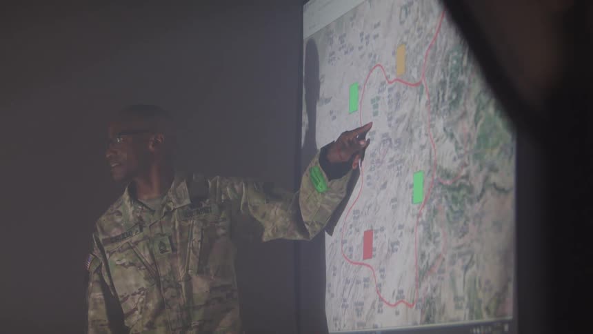 A solider standing in front of a projector screen of a map and pointing and describing the route on the map