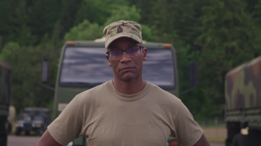 A man in a military uniform stands and poses in front of several military transportation trucks