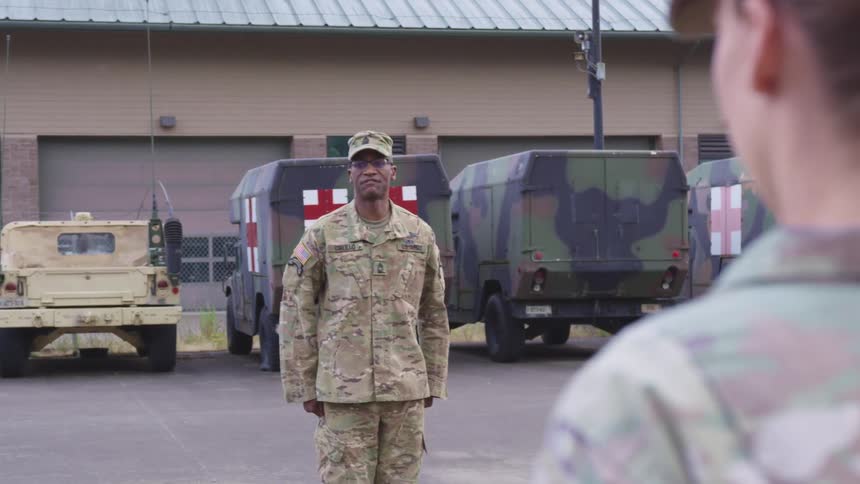 A drill instructor issues parade orders to a female soldier in a military parking lot
