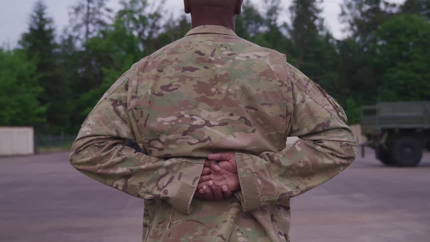 Army soldier standing at ease with hands behind his back in a yard near the treeline