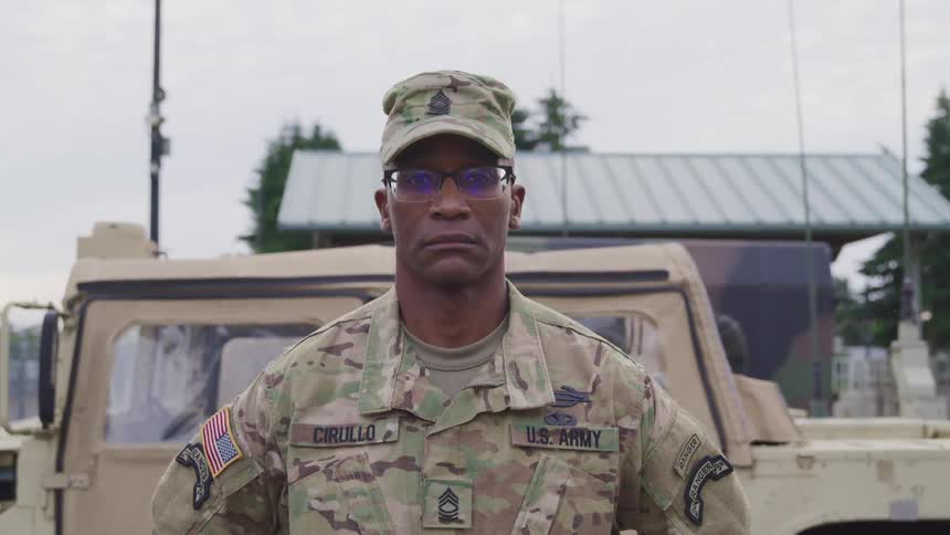 A man in a us army uniform stands with arms behind his back and a stern look on his face