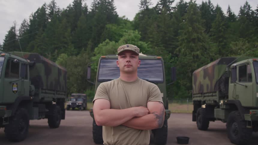 Strong muscular army soldier standing in front of military vehicles parked in a yard looks at the camera