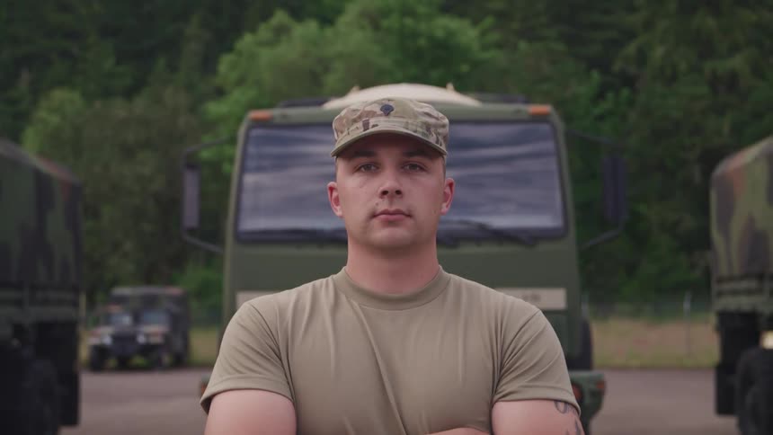 A soldier stands with his arms crossed in front of camouflaged military trucks