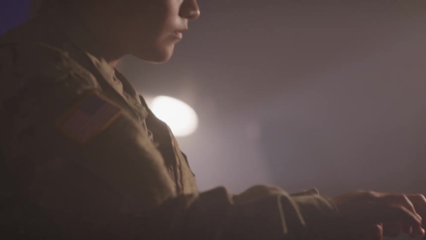 A soldier types on a laptop in a dark room with a lamp