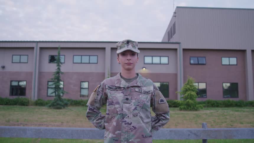 A uniformed female soldier stares into the camera while standing at ease outside of a building