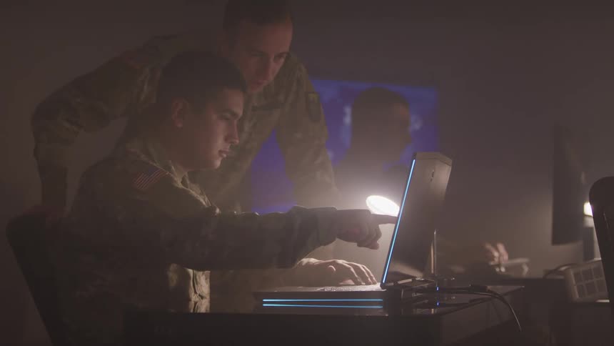 Two soldiers look at a laptop as another types at a computer