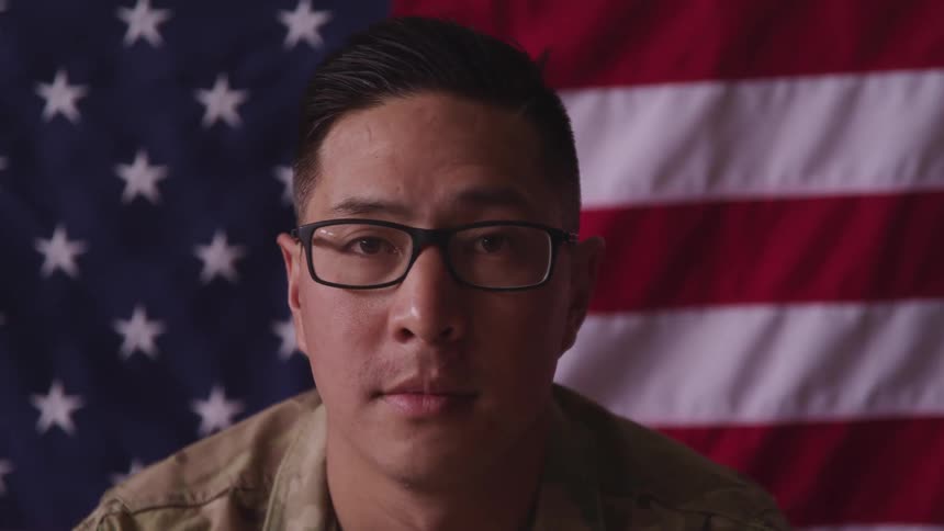 A man with glasses and a military uniform in a room with an american flag in the background