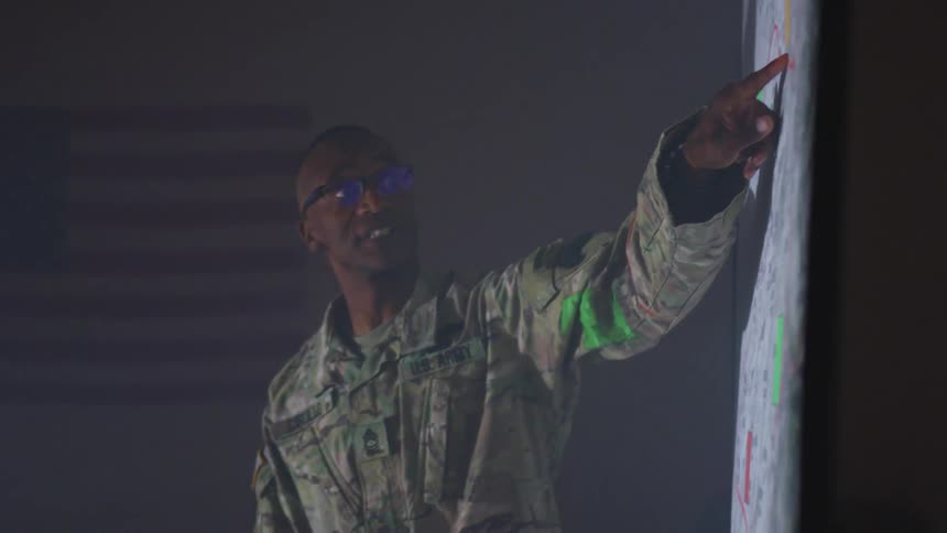 A soldier in camouflage pointing to a map on a wall