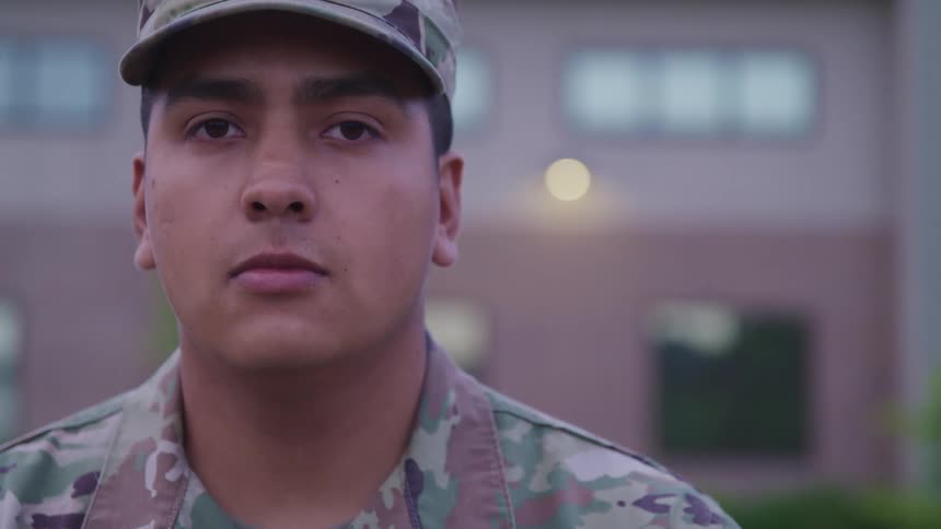 A uniformed soldier stares into the camera while standing outside a building during the day